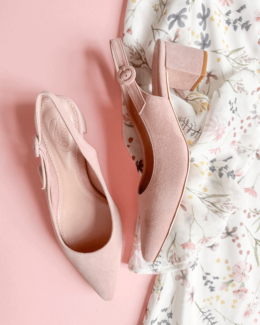 Pink suede slingback heels with a block heel, positioned on a soft pink background. The shoes feature a pointed toe and a delicate bow detail on the side. A floral-patterned fabric is partially visible in the background, adding a feminine touch to the overall presentation. image