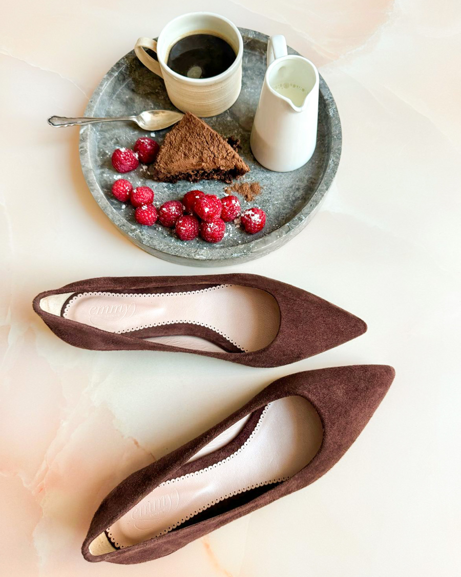 Brown suede pointed-toe heels are positioned on a light marble surface. In the background, a round tray holds a cup of coffee, a small pitcher of cream, a slice of chocolate cake, and fresh raspberries, creating a cozy caf√© setting. The angle captures the shoes from above, emphasizing their sleek design. image