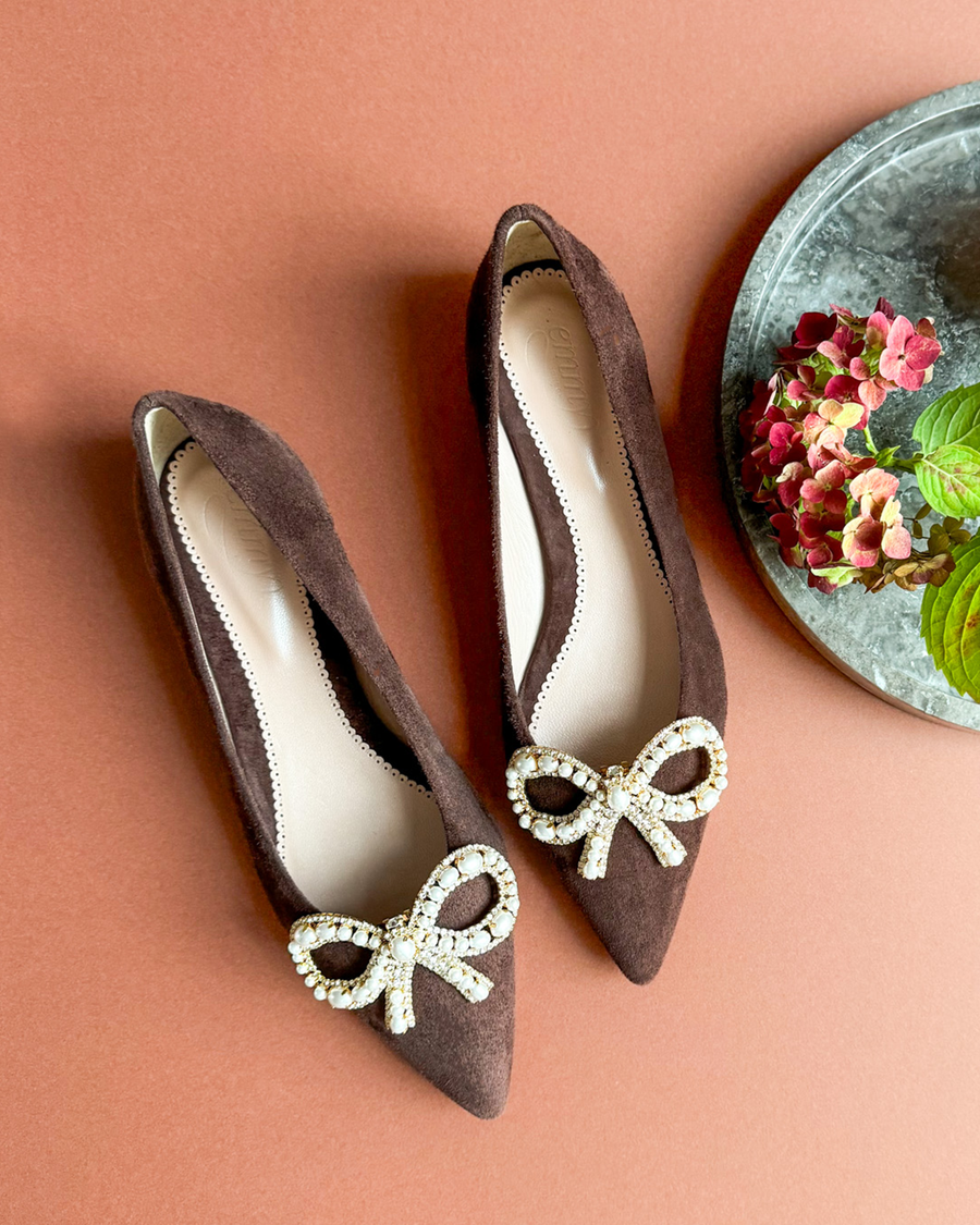 Brown suede heels featuring a decorative bow with rhinestones at the toe. The shoes are positioned on a warm-toned background, with a round stone plate and colorful flowers in the background, showcasing a top-down angle. image