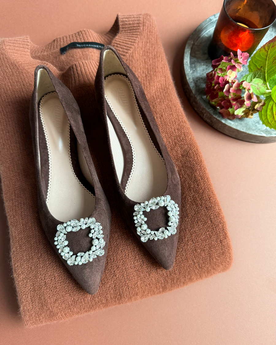 Brown suede heels with a pointed toe and a decorative crystal buckle on the front, placed on a textured rust-colored sweater. The image is taken from a top-down angle, showcasing the shoes against a warm, neutral background with a small vase of flowers and a candle in the corner. image