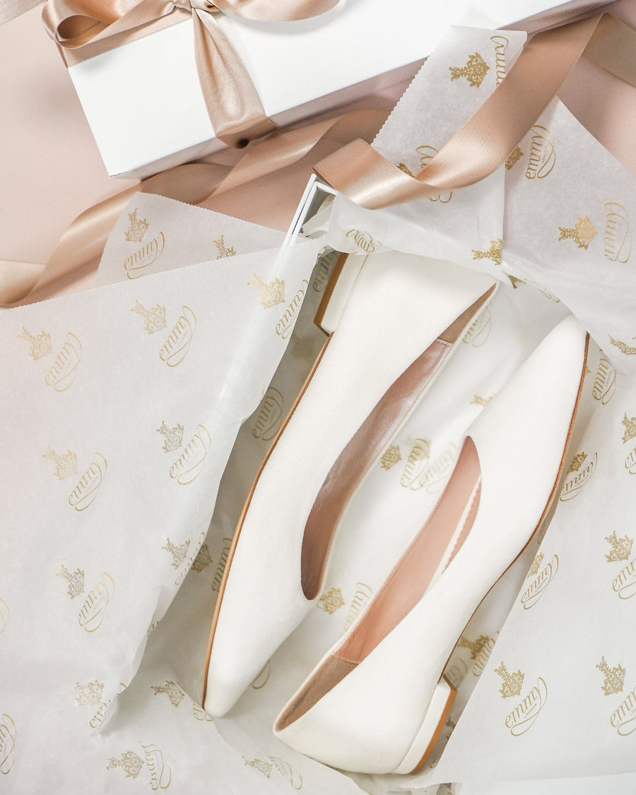 Elegant white shoes are displayed atop a bed of tissue paper featuring a gold crown pattern, surrounded by soft pink wrapping and a ribbon. The shoes have a sleek, pointed design and are positioned at an angle, showcasing their smooth finish and understated elegance. image