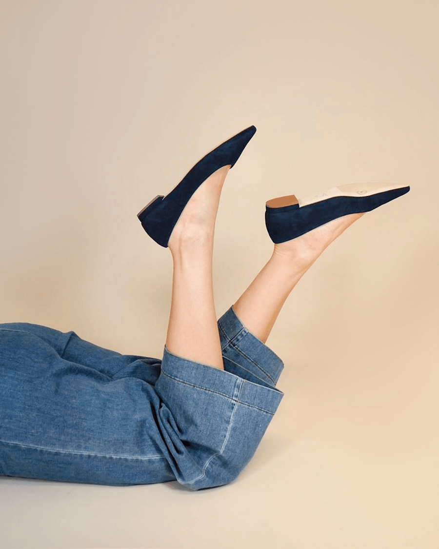 Navy suede heels with a pointed toe, featuring a low block heel. The model is lying on her back with her legs raised, showcasing the shoes against a neutral beige background. The denim pants add a casual touch to the overall look. image
