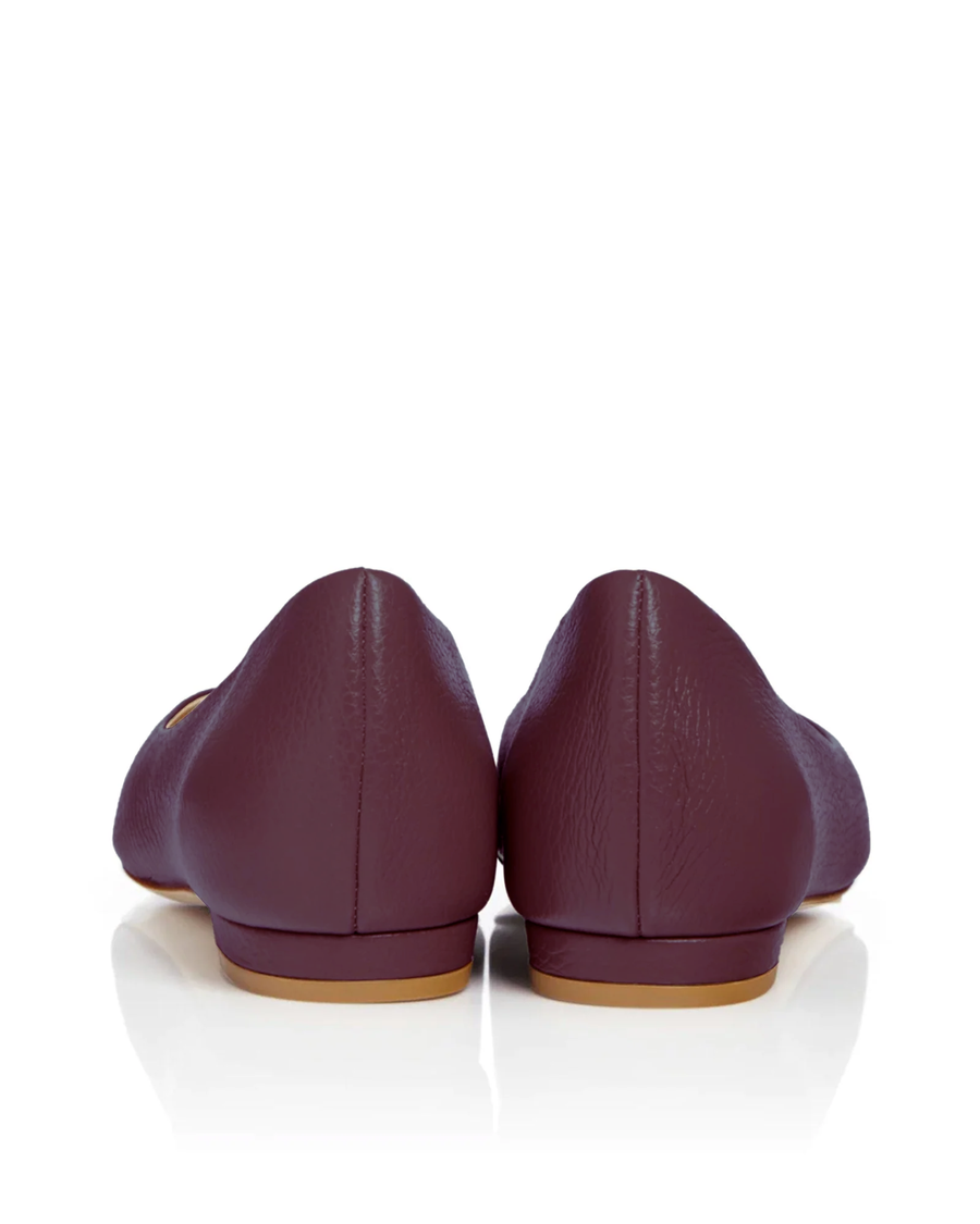 Back view of burgundy flats with a smooth leather finish, showcasing a low heel. The shoes are positioned on a reflective white surface, highlighting their sleek design. The background is plain white, emphasizing the shoes without distractions. image