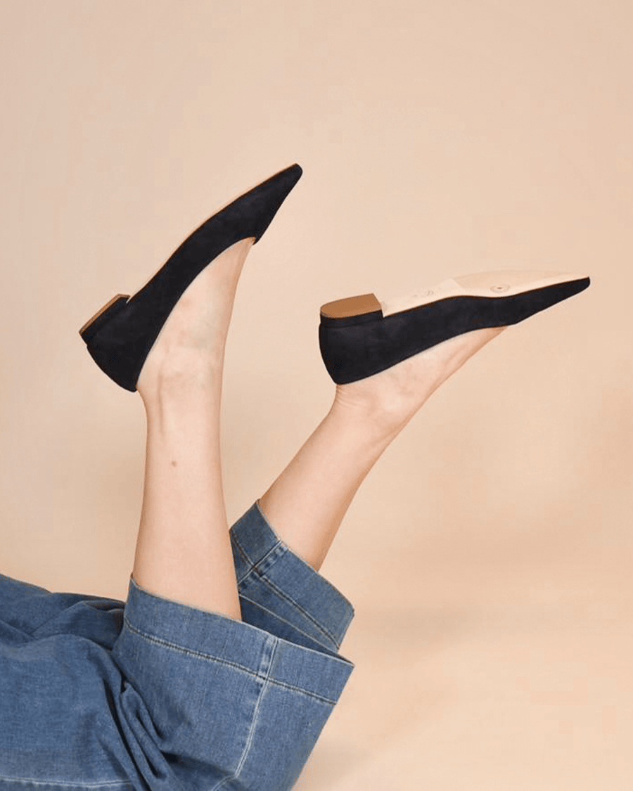 Navy suede flats with a low heel, showcased against a soft beige background. The model is lying down with her legs raised, wearing denim wide-leg pants. The angle highlights the sleek design of the shoes. image