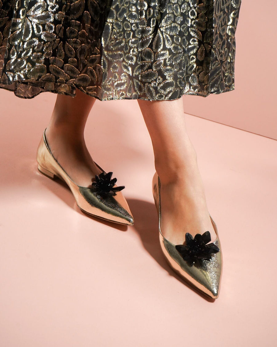 Gold metallic flats with a pointed toe and a decorative black flower embellishment on the front. The model is wearing them with a flowy, patterned skirt, standing on a pink background. The shoes have a flat heel. image
