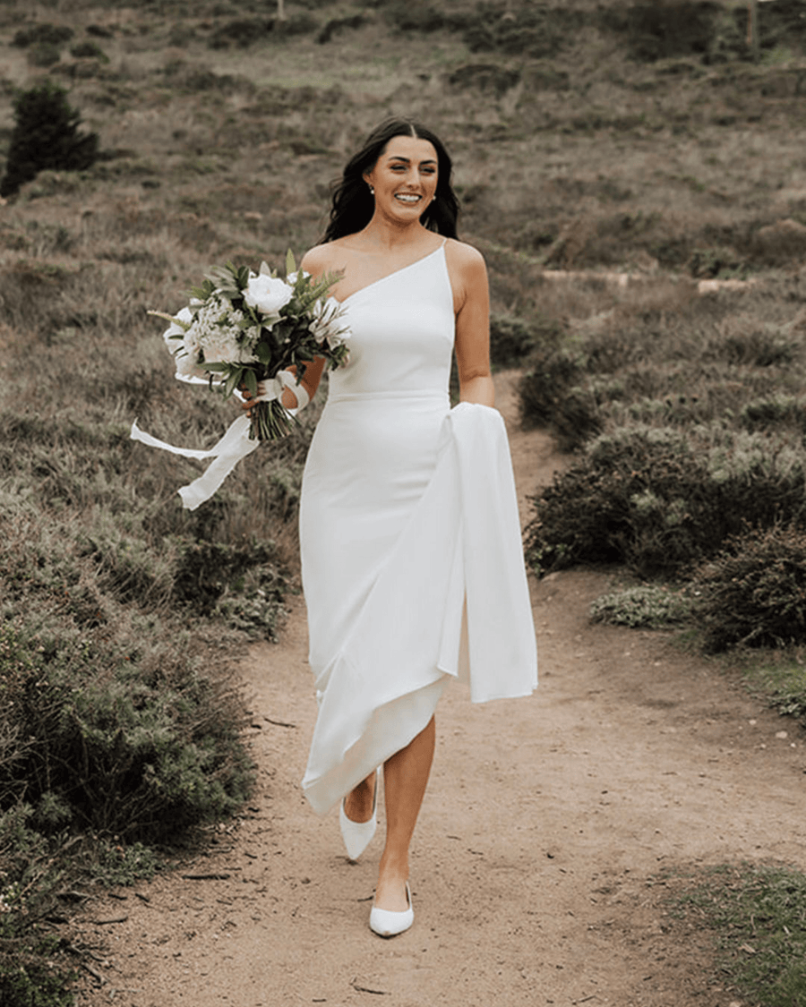 A woman is walking along a dirt path in a natural setting, wearing a sleek, one-shoulder white dress. She holds a bouquet of white flowers and has a white shawl draped over her arm. The shoes are elegant and have a low heel, complementing her outfit. The background features greenery and a soft, earthy landscape. image