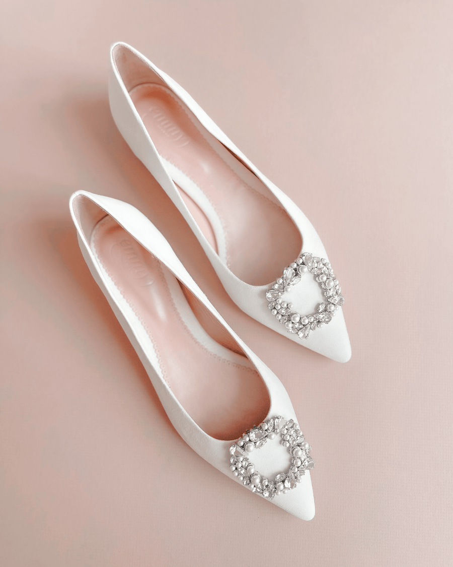 White pointed-toe flats featuring a decorative crystal buckle on the vamp. The shoes are displayed on a soft pink background, showcasing their elegant design. The angle captures both shoes from above, highlighting their sleek silhouette. image
