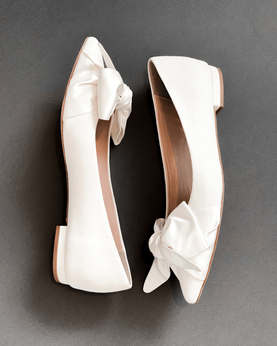 White flat shoes with a bow detail on the toe, set against a dark gray background. The shoes are positioned side by side, showcasing their smooth surface and elegant design. image