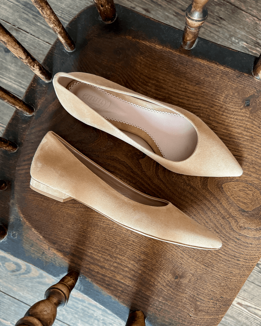 Pointed-toe beige suede heels are displayed on a rustic wooden chair. The shoes are positioned from a top-down angle, showcasing their sleek design and subtle texture. The background features the natural grain of the wood, enhancing the earthy aesthetic. image