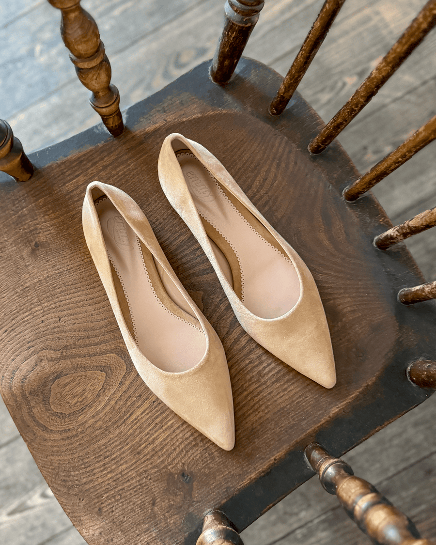 Beige suede pointed-toe flats are displayed on a rustic wooden chair. The shoes are positioned side by side, showcasing their sleek design and smooth surface. The background features a softly lit wooden floor, enhancing the warm tones of the shoes. image
