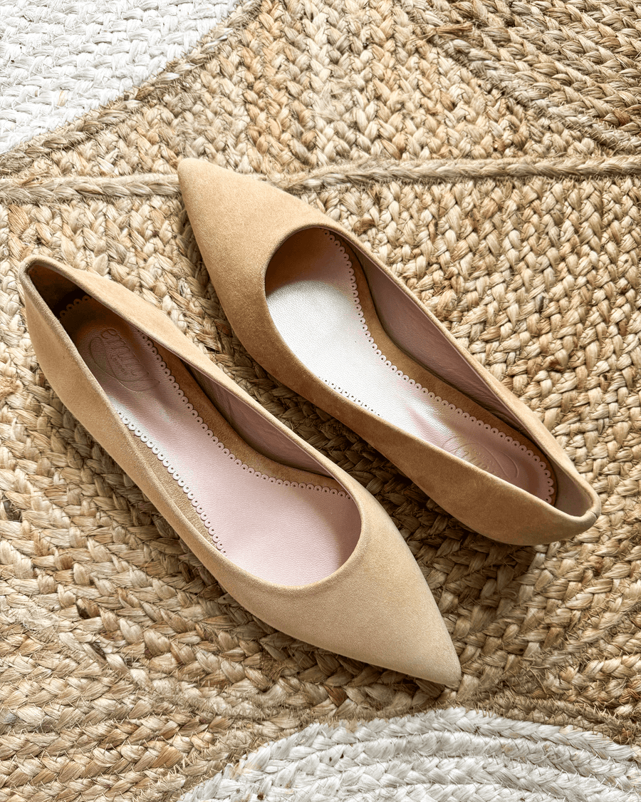 Pointed-toe flats in a soft beige suede material, featuring a cut-out design on the sides. The shoes are positioned on a textured woven background, showcasing their elegant shape and color. image