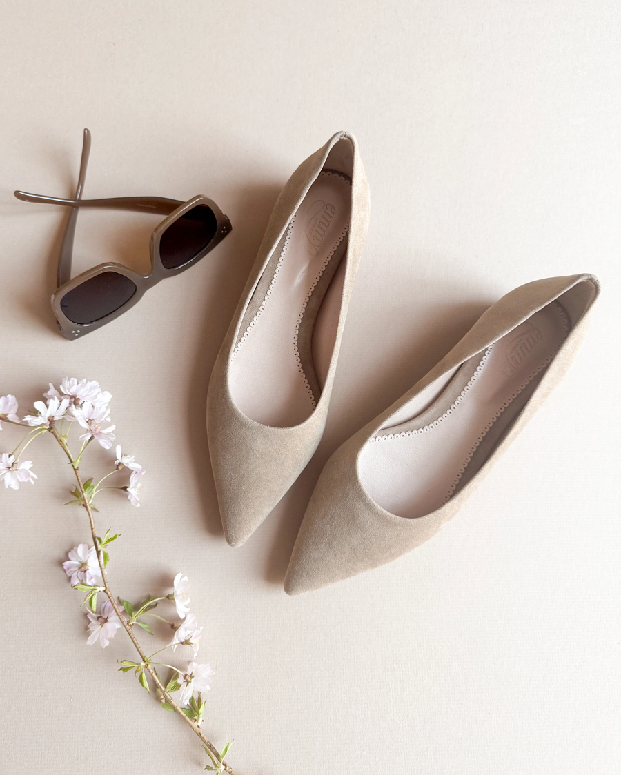 Pointed-toe beige suede flats are displayed on a light beige background, accompanied by a pair of stylish sunglasses and a delicate branch of pink flowers. The shoes are positioned side by side, showcasing their sleek design and soft texture. image