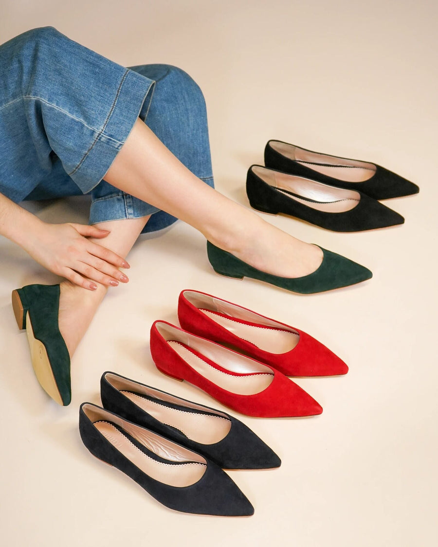 A model is seated on the floor, wearing denim culottes, with her legs crossed. She is showcasing three pairs of pointed-toe pumps in red, green, and black suede, arranged neatly in front of her. The background is a soft beige, enhancing the colors of the shoes. The heel height is approximately 2 inches. image