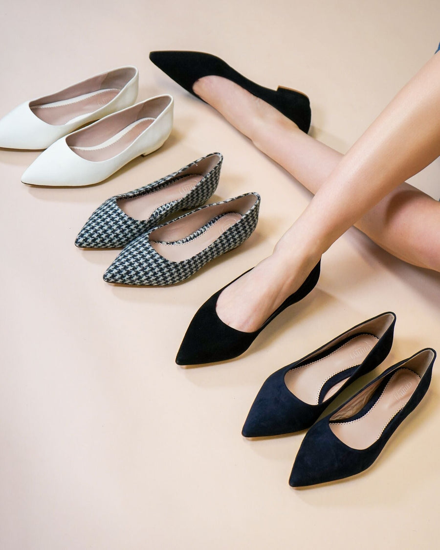 A collection of three pairs of pointed-toe shoes displayed on a light beige background. The first pair is white leather flats, the second pair features a black and white tweed pattern, and the third pair is black suede. A model's legs are visible, wearing the black suede shoes, showcasing the elegant design and fit. image