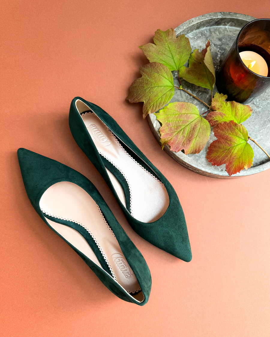 Green suede pointed-toe heels are displayed on a warm orange background, accompanied by a round marble tray featuring autumn leaves and a lit candle. The shoes are positioned from a top-down angle, showcasing their elegant design and the delicate scalloped edge of the insole. image