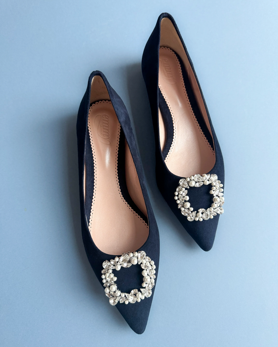 Elegant navy suede flats featuring a striking embellished buckle at the toe. The shoes are positioned on a soft blue background, showcasing their pointed toe design. The image captures the shoes from a top-down angle, highlighting the smooth interior and the intricate detailing of the buckle. image