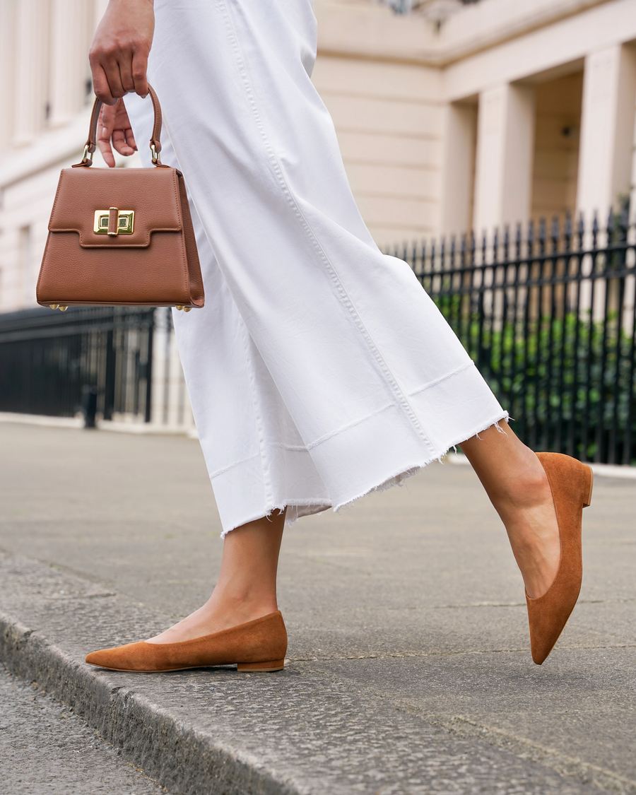 A woman is walking on a city street, wearing a pair of brown suede flats. The shoes have a pointed toe and a low profile, complementing her white, ankle-length, frayed hem pants. She carries a small brown handbag, and the background features a classic building with a black railing. The angle captures her side profile, emphasizing the stylish combination of her outfit. image
