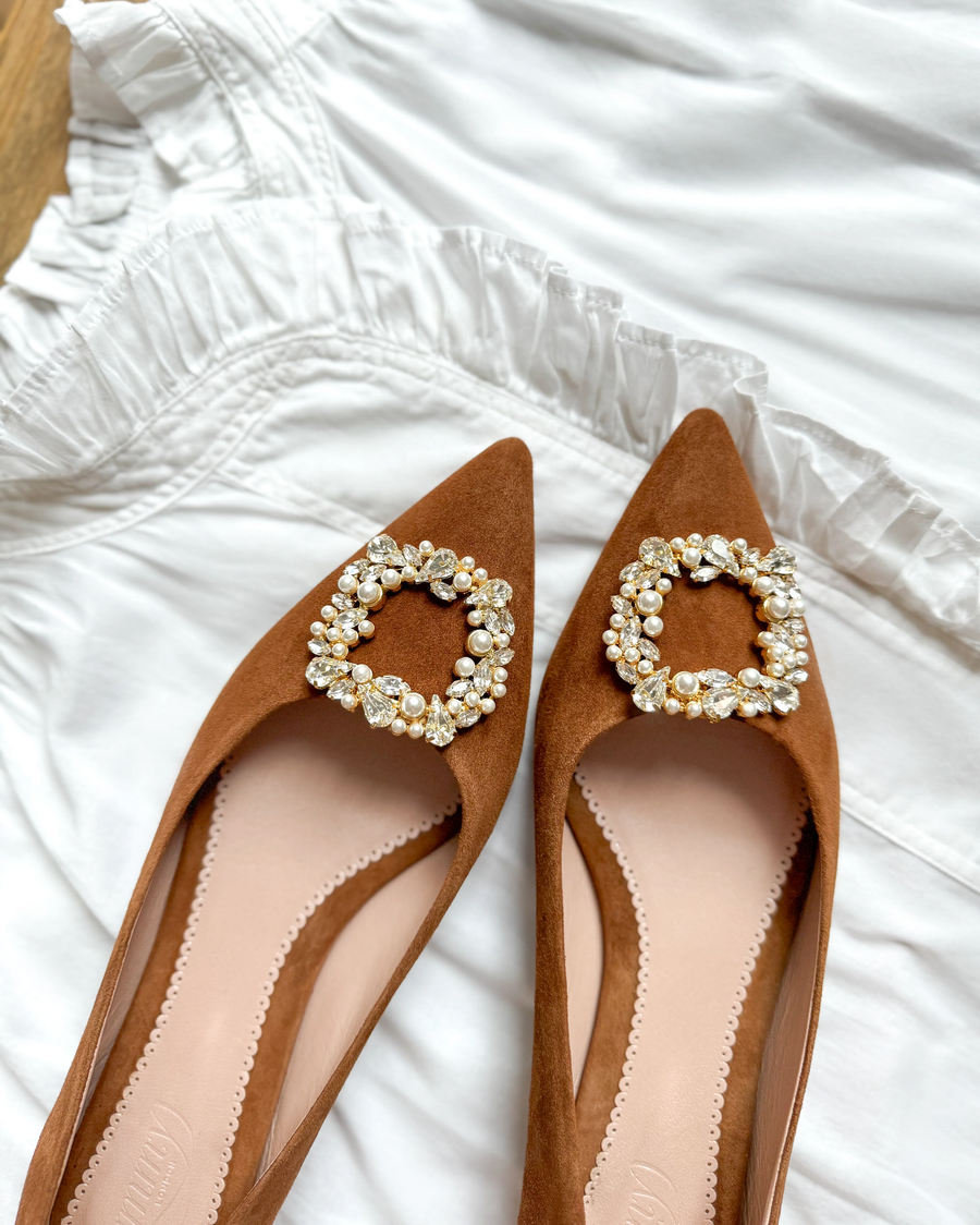 Brown suede pointed-toe flats featuring a decorative pearl buckle on the vamp, set against a white ruffled dress. The shoes are positioned at an angle, showcasing the intricate detailing of the buckle and the soft texture of the suede. image
