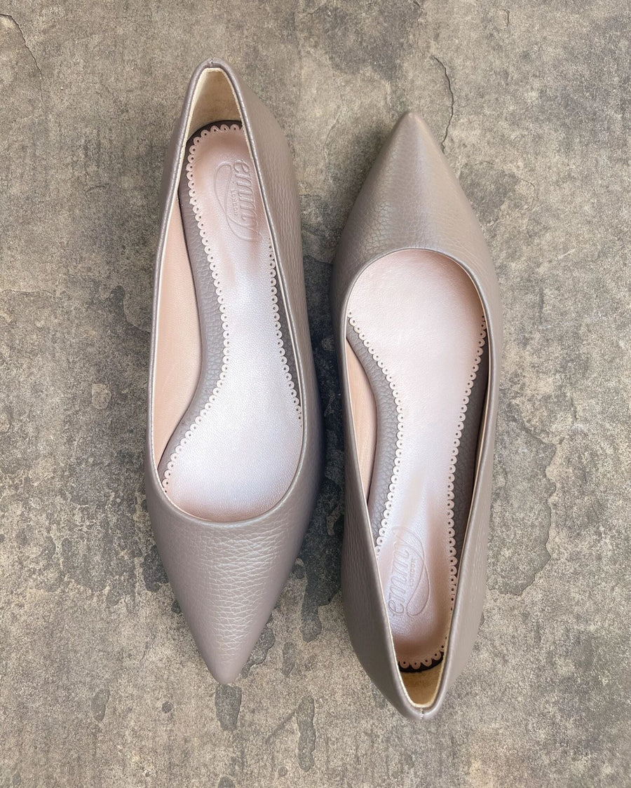 Pointed-toe shoes in a soft taupe leather, featuring a sleek design with a smooth interior lining. The shoes are displayed from a top-down angle on a textured gray surface, showcasing their elegant silhouette. image