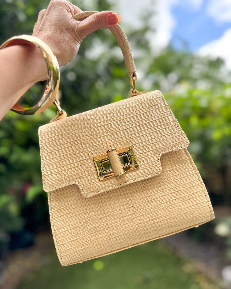 Beige handbag with gold accents held by a hand against a blurred green background image
