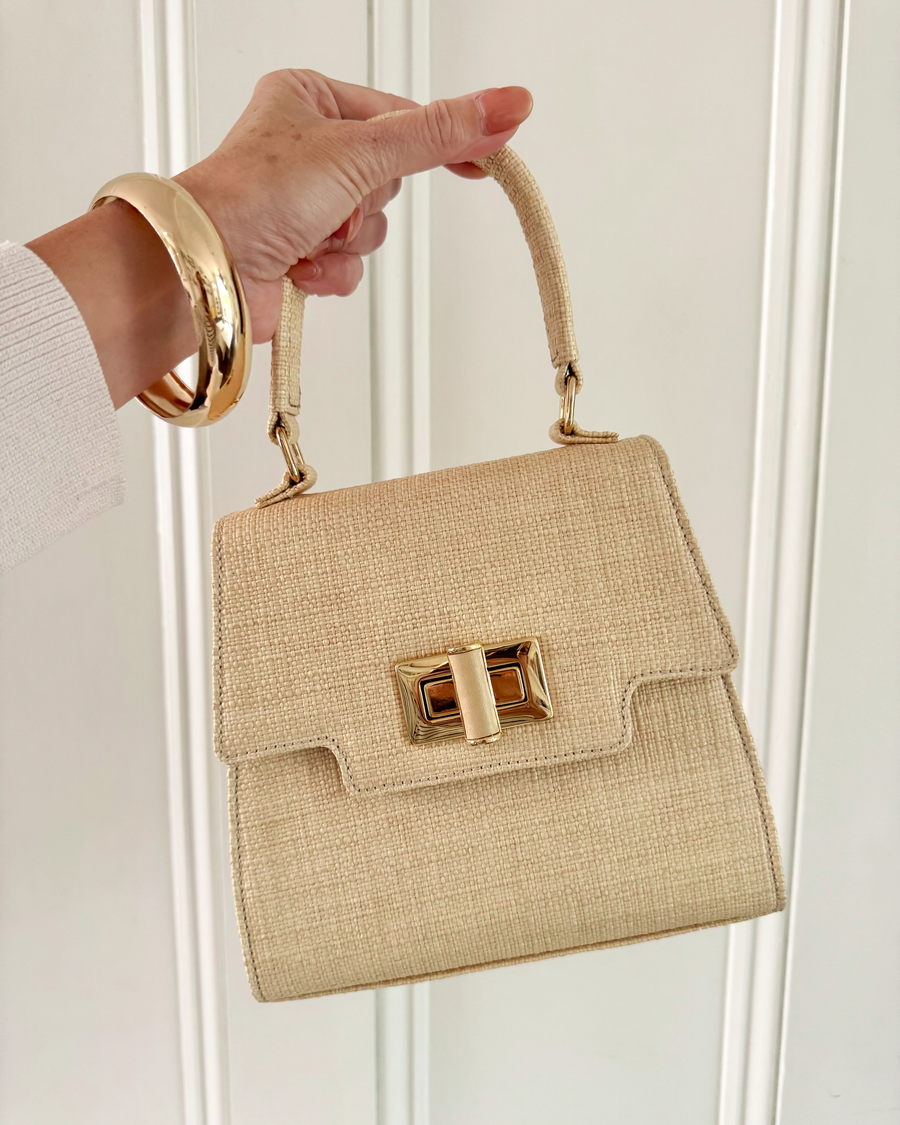Beige handbag with gold accents held by a hand against a white background image