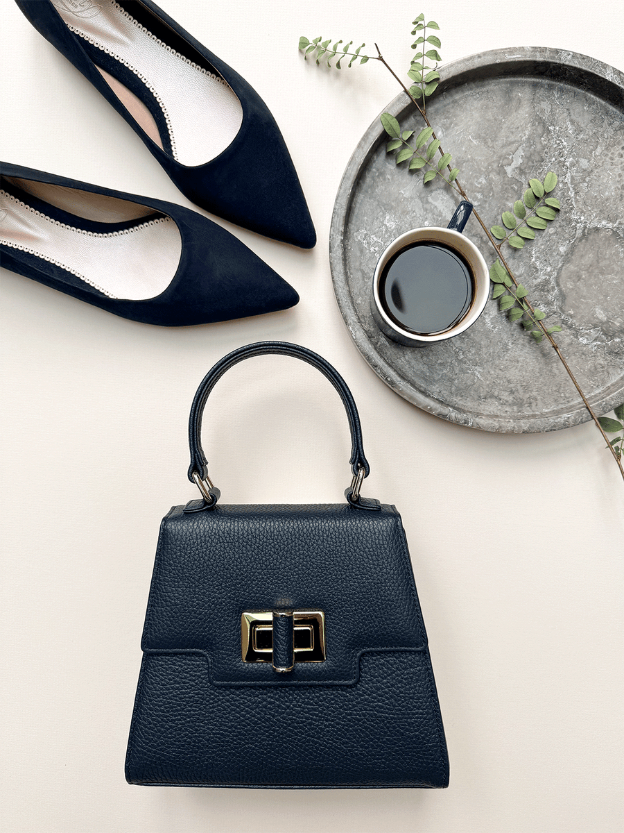 Navy suede pointed-toe flats are positioned above a round silver tray with a small cup of coffee and a sprig of greenery. The image is taken from a top-down angle, showcasing the shoes alongside a stylish black handbag with a gold clasp. The background is a soft, neutral tone, enhancing the elegance of the arrangement. image