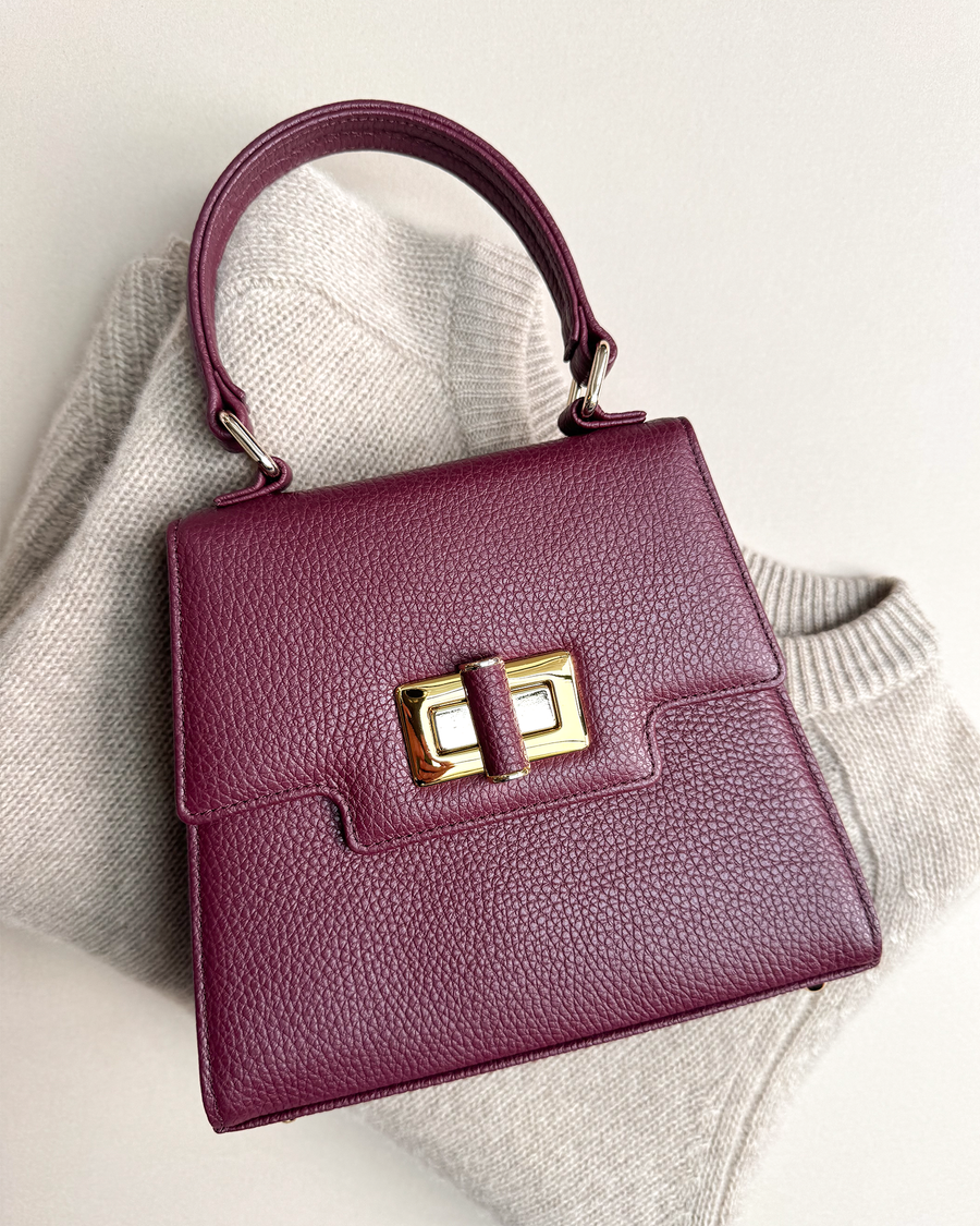 A deep burgundy handbag with a structured design sits atop a light beige knitted sweater. The bag features a gold-tone clasp and a top handle, showcasing a sophisticated and elegant style. The background is neutral, emphasizing the rich color of the handbag. image