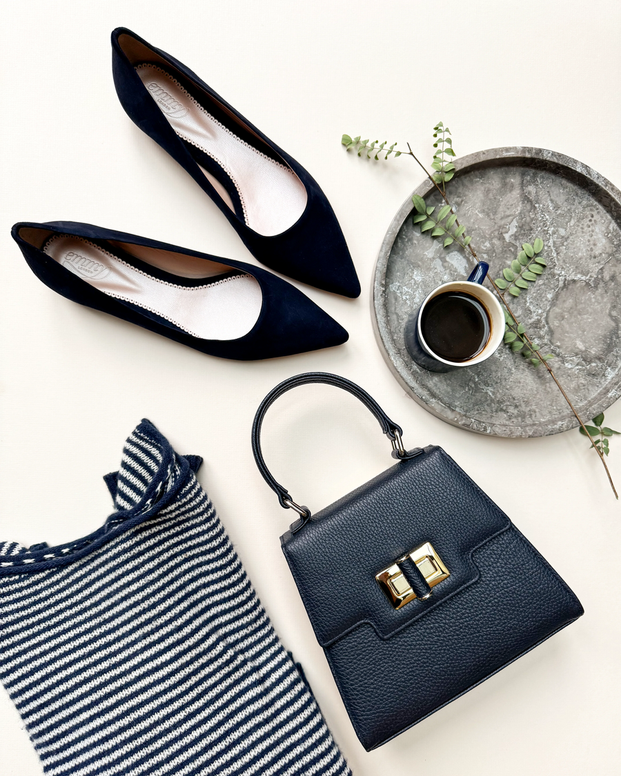 Navy suede pointed-toe heels are positioned alongside a stylish black handbag and a small cup of coffee on a light-colored surface. A striped fabric is partially visible in the foreground, adding texture to the arrangement. The image is taken from a top-down angle, showcasing the elegant design of the shoes and accessories. image