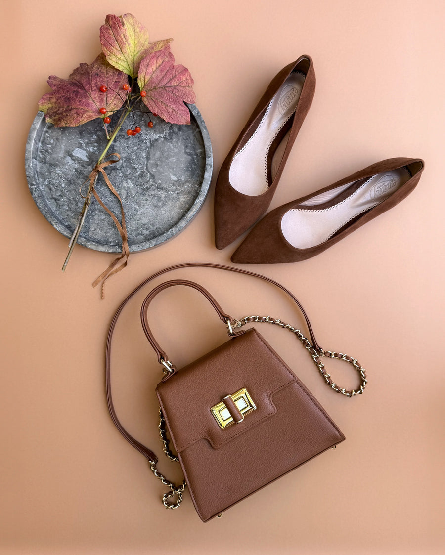 Brown suede pointed-toe heels are positioned next to a stylish brown handbag on a soft peach background. The heels feature a sleek design, showcasing a moderate heel height. A decorative arrangement of autumn leaves adds a touch of seasonal charm to the composition. image
