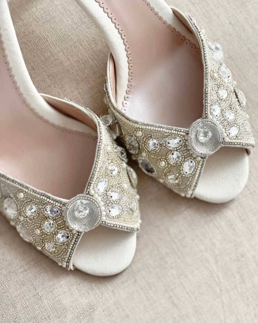 Elegant open-toe heels featuring intricate embellishments with rhinestones and floral accents. The shoes are set against a soft, neutral background, showcasing their delicate design. The angle highlights the detailed craftsmanship and the luxurious feel of the materials used. image