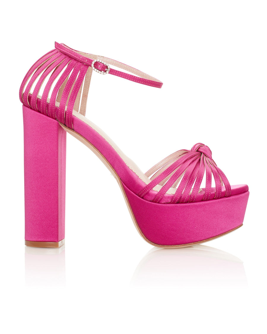 Strappy pink platform heels with a chunky block heel. The shoes feature a wide ankle strap and a knotted front design. The image is taken from a side angle against a white background, showcasing the vibrant color and sleek silhouette. image