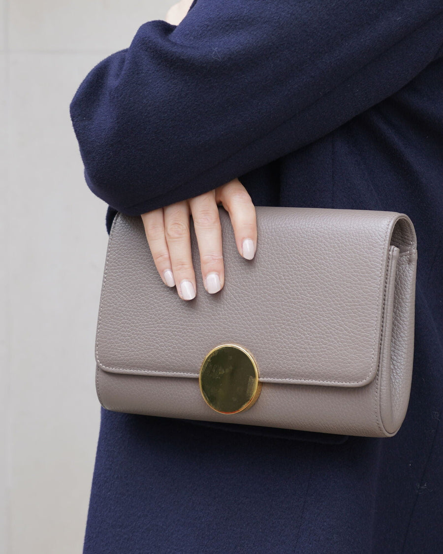 A close-up image of a person holding a taupe clutch bag with a circular gold clasp. The bag is made of leather and features a smooth texture. The individual is wearing a dark navy coat, and the background is a neutral, light-colored wall. The angle of the image focuses on the bag and the person's hand, showcasing the accessory prominently. image