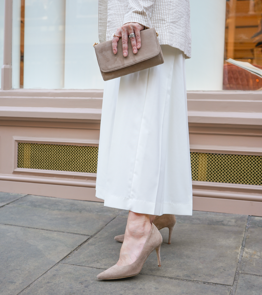 A woman is standing on a stone pavement, wearing a long white skirt and a light-colored blazer. She holds a beige clutch in her right hand. The shoes are pointed-toe heels made of suede, featuring a stiletto heel that is approximately 4 inches high. The background shows a stylish storefront with large windows. image