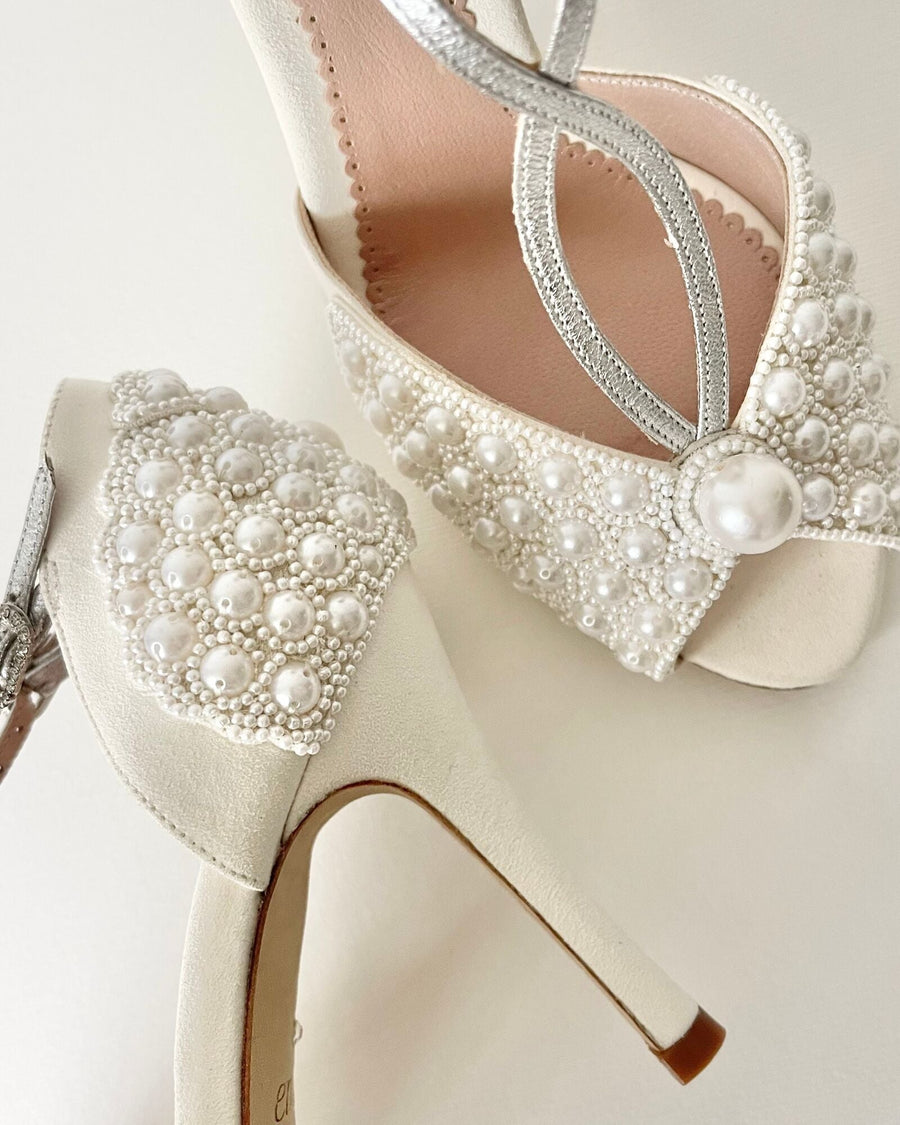 Elegant white heels featuring intricate pearl embellishments on the toe area and a delicate silver strap. The shoes are positioned on a light-colored surface, showcasing their refined design. The heel height is approximately 4 inches, and the material is likely leather. image