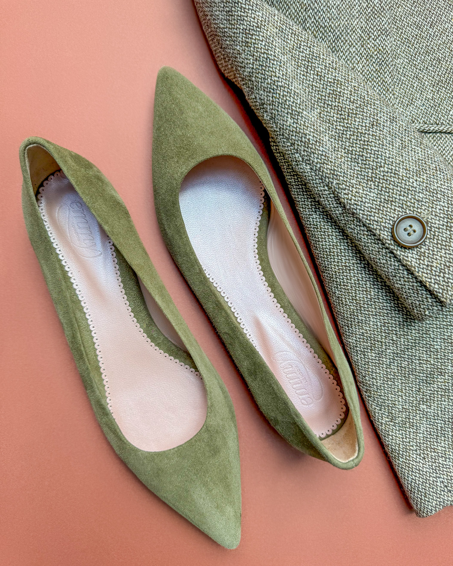 Green suede pointed-toe flats are displayed on a coral background, alongside a textured tweed blazer. The shoes feature a sleek design with a low heel, emphasizing a chic and modern aesthetic. The angle captures both the shoes and the blazer, showcasing their complementary styles. image