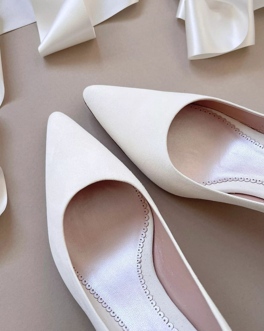 White pointed-toe heels with a smooth finish, set against a neutral beige background. The shoes feature a sleek design with a cut-out detail on the sides, showcasing a soft interior. The image is taken from a top-down angle, highlighting the elegant shape and clean lines of the footwear. image