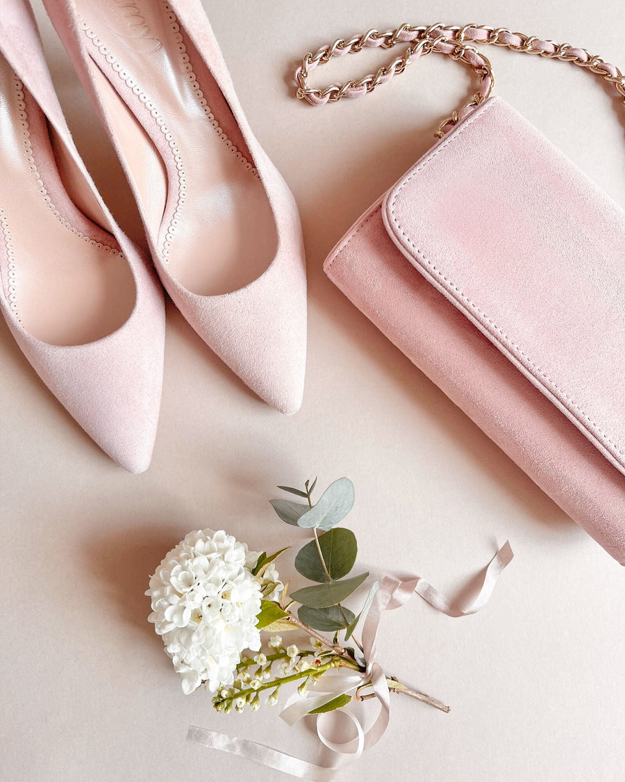 Pink suede heels with a pointed toe are positioned next to a matching pink suede clutch. A small bouquet of white flowers and greenery, along with a delicate ribbon, are arranged nearby on a soft beige background. The image is taken from a top-down angle, showcasing the elegant pairing of the shoes and bag. image