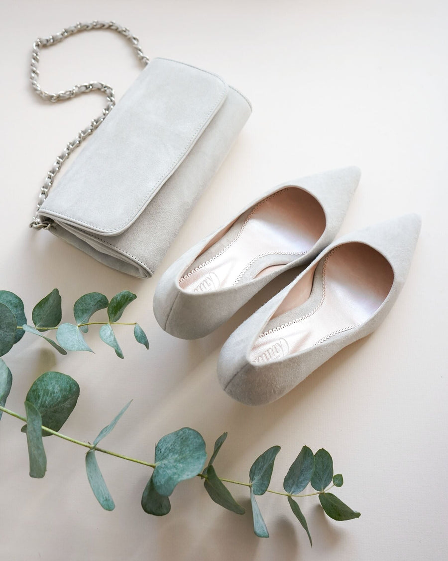 Light gray suede pointed-toe heels are displayed alongside a matching gray clutch purse. The shoes are positioned on a neutral background with a sprig of eucalyptus nearby, showcasing a flat lay angle. The heel height appears to be mid. image