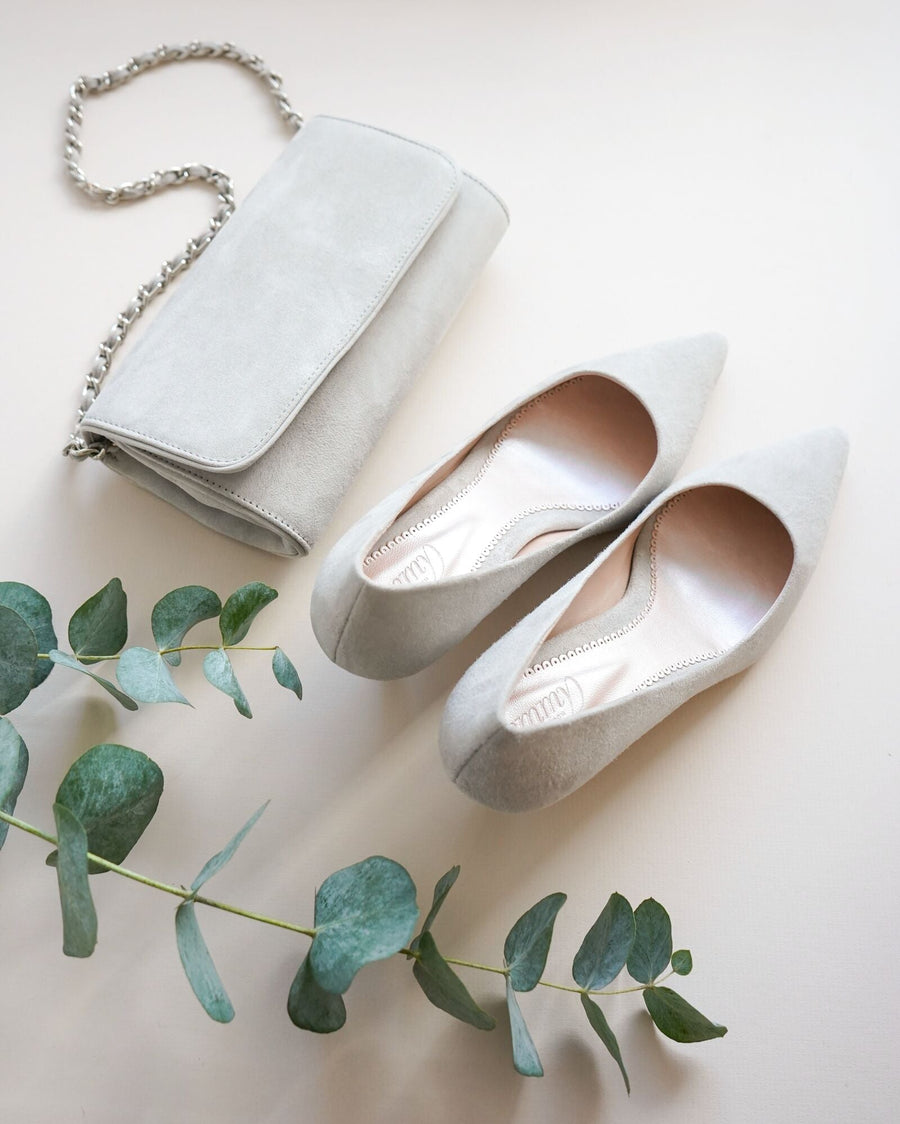 Pointed-toe suede heels in a light gray shade are displayed alongside a matching gray clutch purse. The shoes are positioned on a soft, neutral background with a sprig of eucalyptus nearby, creating a serene and elegant composition. The angle showcases the shoes from above, highlighting their sleek design and the smooth interior. image