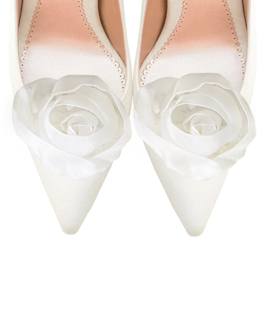 Elegant white pointed-toe heels featuring a delicate fabric rose embellishment on the front. The shoes are set against a clean, neutral background, showcasing their refined design. The angle highlights the intricate detailing of the rose and the smooth finish of the shoe. image