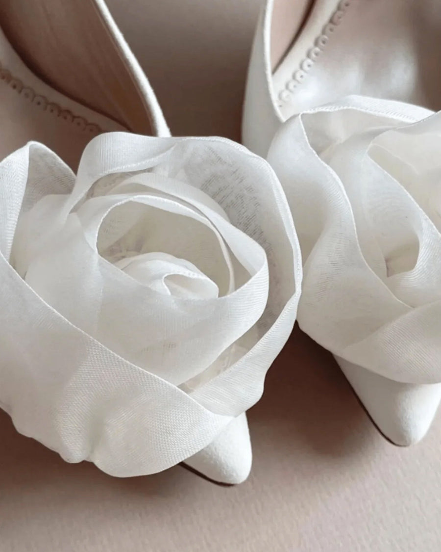 Elegant white pointed-toe heels adorned with delicate sheer fabric roses at the front. The shoes are positioned closely together, showcasing the intricate floral details. The background is softly blurred, emphasizing the shoes. The angle captures the front view, highlighting the unique design. image