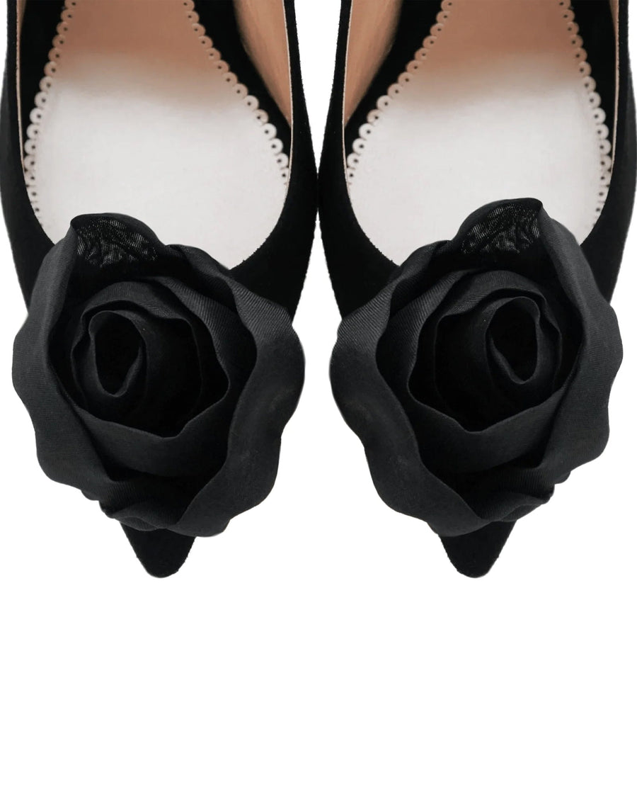 Black pointed-toe heels featuring a large black fabric rose embellishment at the front. The shoes are set against a neutral background, showcasing their elegant design. The angle captures the shoes from above, emphasizing the floral detail. image