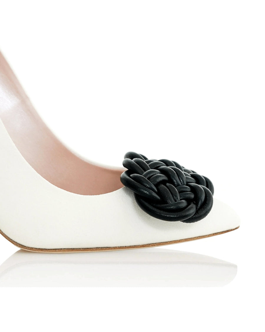 White leather heels featuring a prominent black woven embellishment at the toe. The image is taken from a side angle, showcasing the sleek design and pointed toe. The background is a clean, white surface, emphasizing the shoe's details. image
