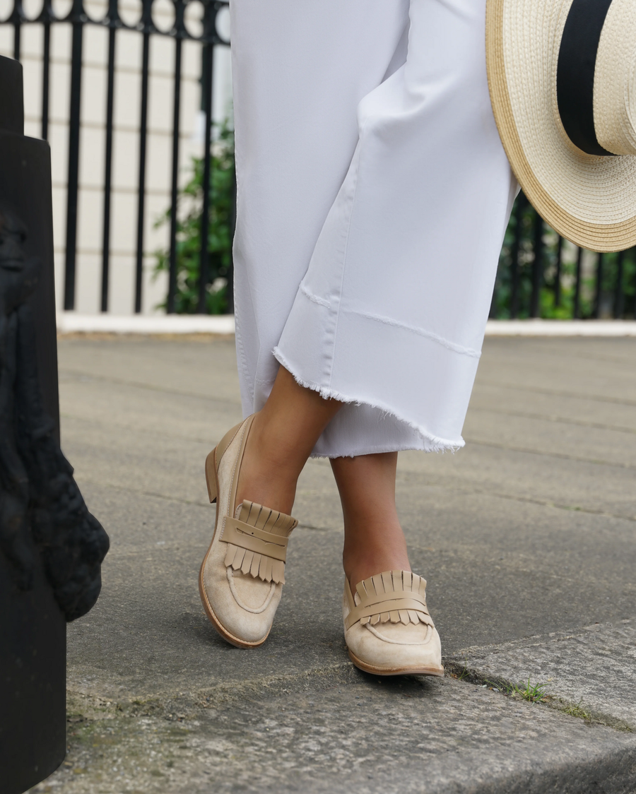 Fringe loafers in a light beige color, made of suede, feature a low heel. The model is standing on a stone pathway, wearing white, cropped pants that have a frayed hem. A wide-brimmed straw hat is partially visible in the background, adding a summery vibe to the scene. image