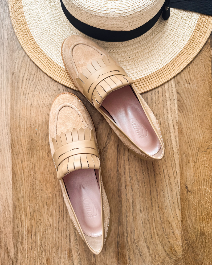Tan loafers made of suede feature a fringed design on the front. The shoes are positioned on a wooden surface, with a straw hat partially visible in the background, creating a casual summer vibe. The angle captures the shoes from above, showcasing their sleek silhouette. image
