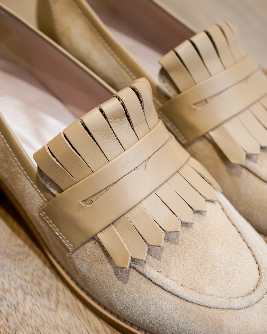Beige loafers featuring a decorative fringe detail on the upper. The shoes are made of a soft material, with a close-up view highlighting the intricate design. The background is a light wooden surface, providing a warm contrast to the shoes. image