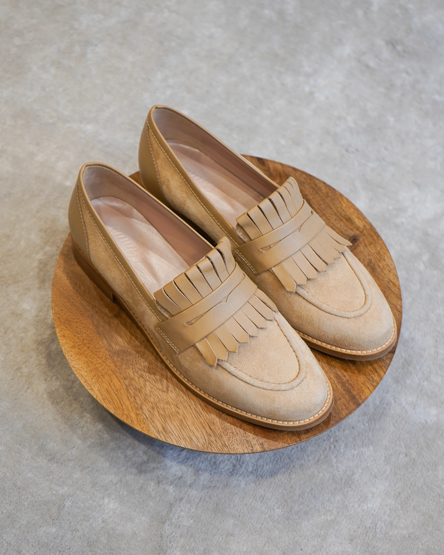 Tan suede loafers with a fringe detail are displayed on a round wooden platform. The shoes have a low heel and are positioned against a neutral gray background, showcasing their elegant design. image