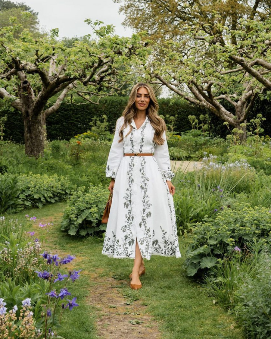 A woman is walking through a lush garden filled with blooming flowers and greenery. She is wearing a long white dress with floral patterns and a brown belt at the waist. The dress has long sleeves and a front slit. She is carrying a brown handbag and wearing tan heels. The image is taken from a frontal angle, showcasing the vibrant outdoor setting. image