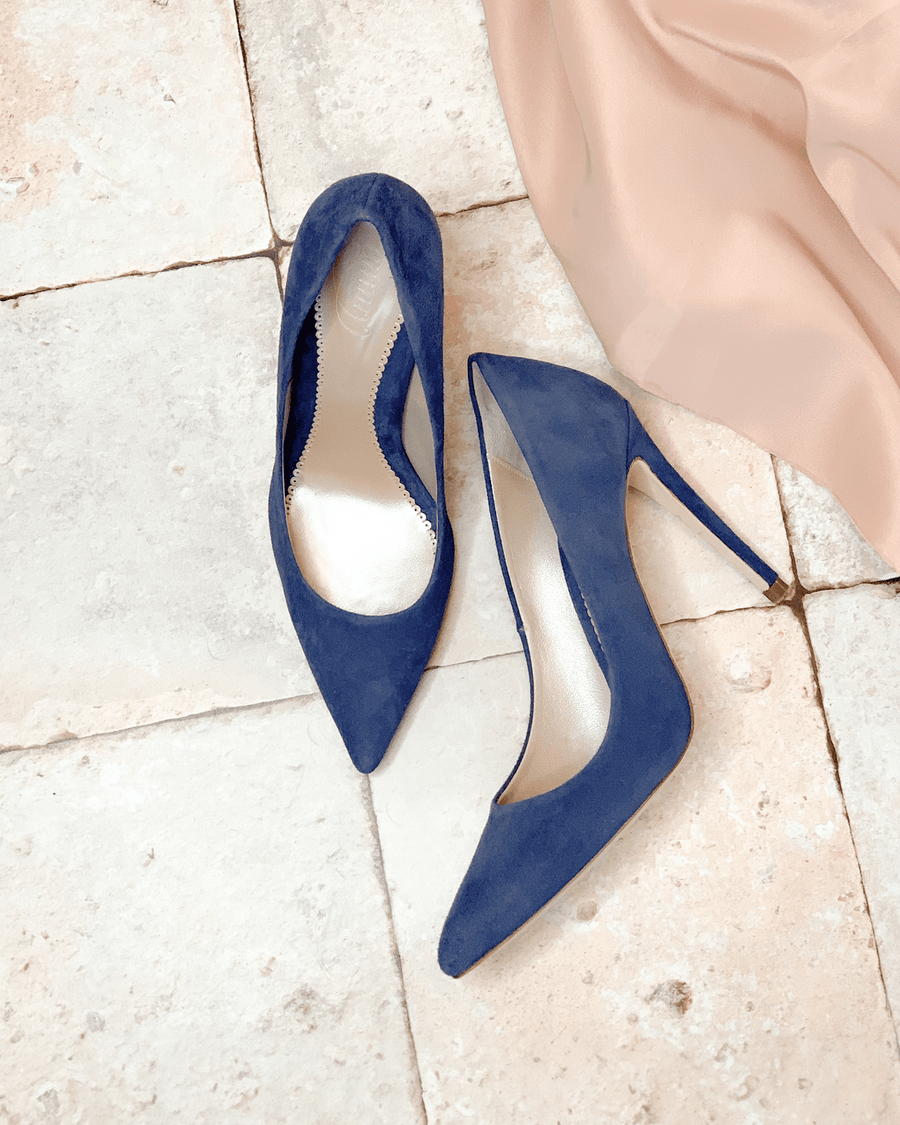 Blue suede high heels with a pointed toe, placed on a light stone floor. A soft, flowing beige fabric is draped nearby, adding a touch of elegance to the scene. image
