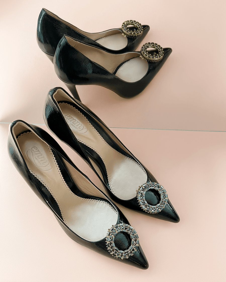 Black patent leather heels with a pointed toe and decorative brooch detail on the front. The shoes are positioned on a soft pink background, showcasing their sleek design. The angle captures both the side and top view of the heels, emphasizing their elegant silhouette. image